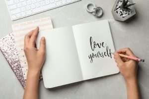 Woman writing LOVE YOURSELF in journal on grey table, codependency recovery in Florida | Codependency Treatment | online therapy in Florida | Counseling Solutions of Broward 33023