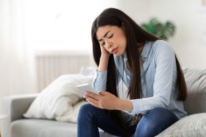Woman sitting on couch, head in her hand looking at her phone upset. She could use online therapy for codependent relationship in florida and post-divorce counseling in florida to help with her codependent coping. Get online counseling in Florida here with Broward counselor, Enid.