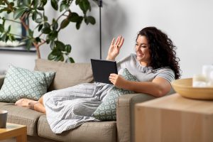 Woman sitting on a couch, holding a tablet and smiling and waving to start anxiety treatment with online therapist in Florida, Enid, for online therapy in Florida. You can get help with imposter syndrome, codependency treatment and perfectionism counseling for women in miramar, fl.