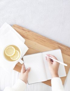 Woman's hand writing in a notebook with a cup and a couple of other notebooks on a white table. She is enjoying the benefits of journaling for high-performing women for confidence building in Miramar, FL. She gets online therapy in Florida for women with anxiety and codependency and perfectionism.